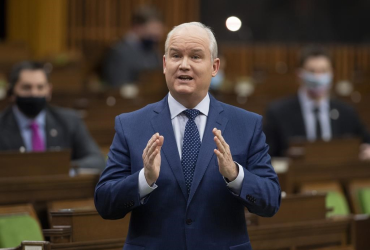 A man in a suit and tie stands and speaks, gesturing with his hands in front of his chest