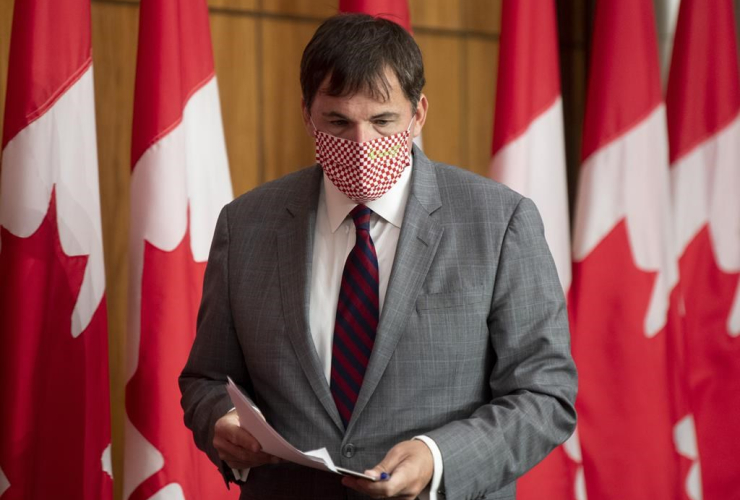 A man in a mask holding papers walks in front of a row of Canadian flags