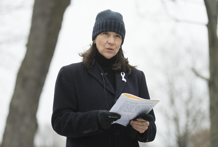 A woman wearing a toque and an overcoat stands outside, reciting a poem from a notebook 