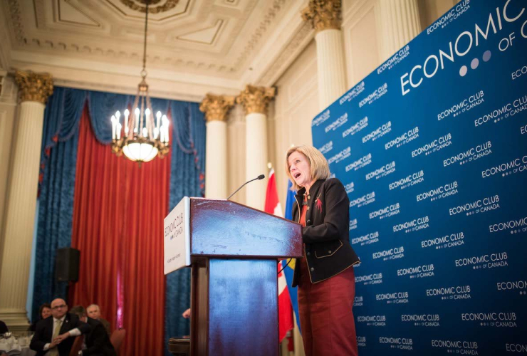 A blond woman in a red dress and black jacket stands behind a podium, speaking, with a large chandelier to her right and a group of people looking on