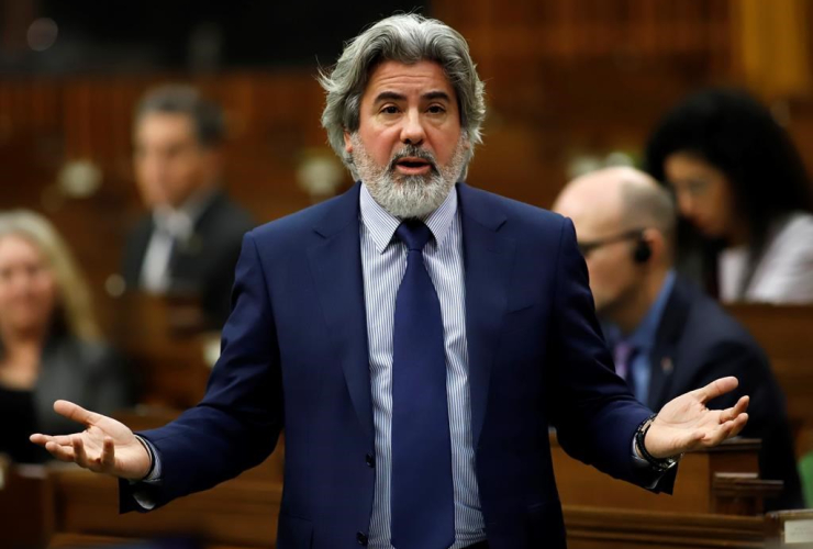 A bearded man with grey hair speaks. He's wearing a navy suit and tie.