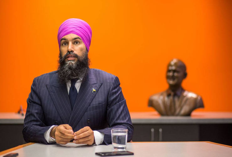 A man in a suit and turban sits in front of a bright orange wall