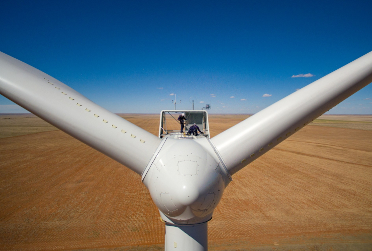 two workers stand atop a huge windmill overlooking a great expanse of field.