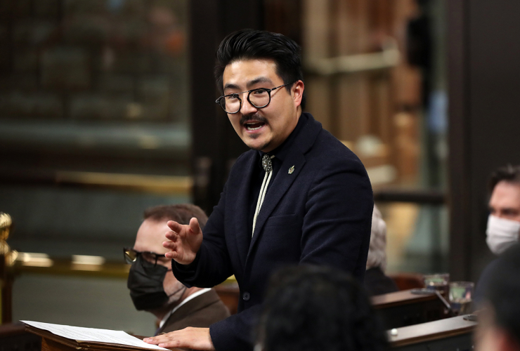 A man wearing a black suit jacket and glasses stands in the House of Commons, gesturing, mid-sentence as his colleagues look on in the background and foreground. 