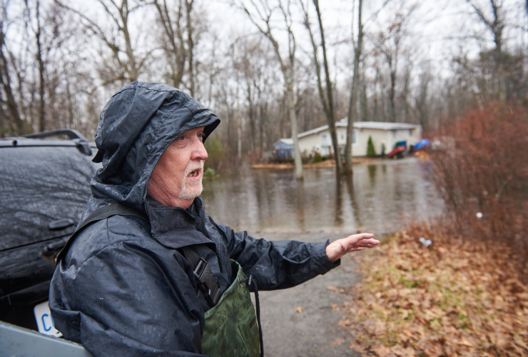 An older man stands in a black raincoat and gestures, with a great expanse of flooded land visible in the background.