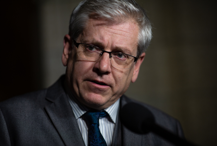 A man with grey hair wearing glasses and a suit looks into the camera.