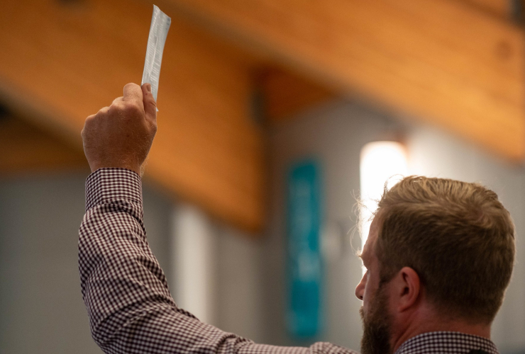 A man in a plaid shirt raises his arm while holding a small piece of paper, face moslty turned away from the camera