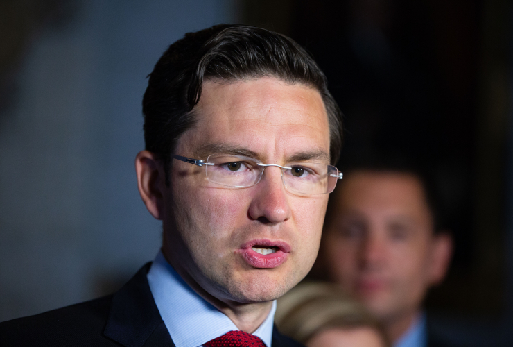 A bespectacled man wearing a suit is photographed mid-sentence