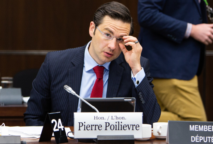 A man in a suit adjust his glasses in Canada's House of Commons, while sitting at a desk