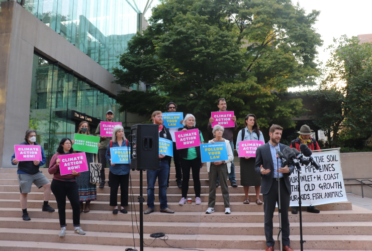 13 people holding signs that say "Climate Now" and "follow your law" stand outside on the steps of a building