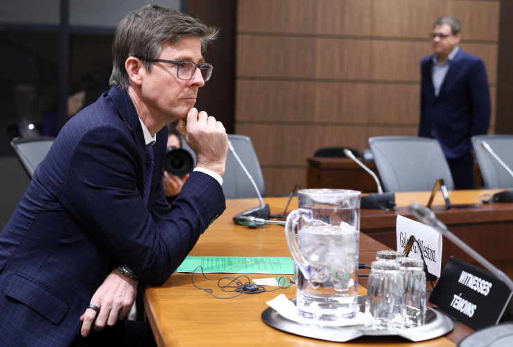 a man wearing glasses and a blue suit sits at a long wooden table, leaning forward on his arms