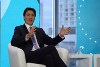 The prime minister of canada (a white man) sits in a white armchair, shown from the shin up, gesturing with both hands