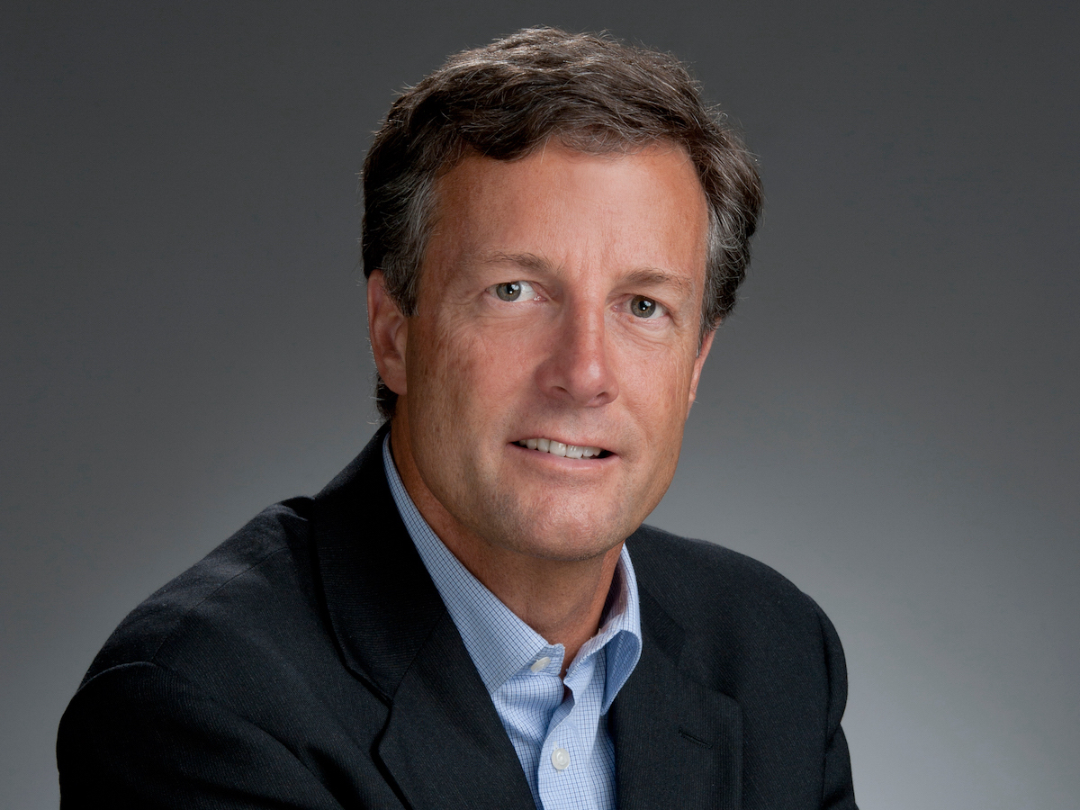 A man with dark brown hair and a suit jacket looks at the camera