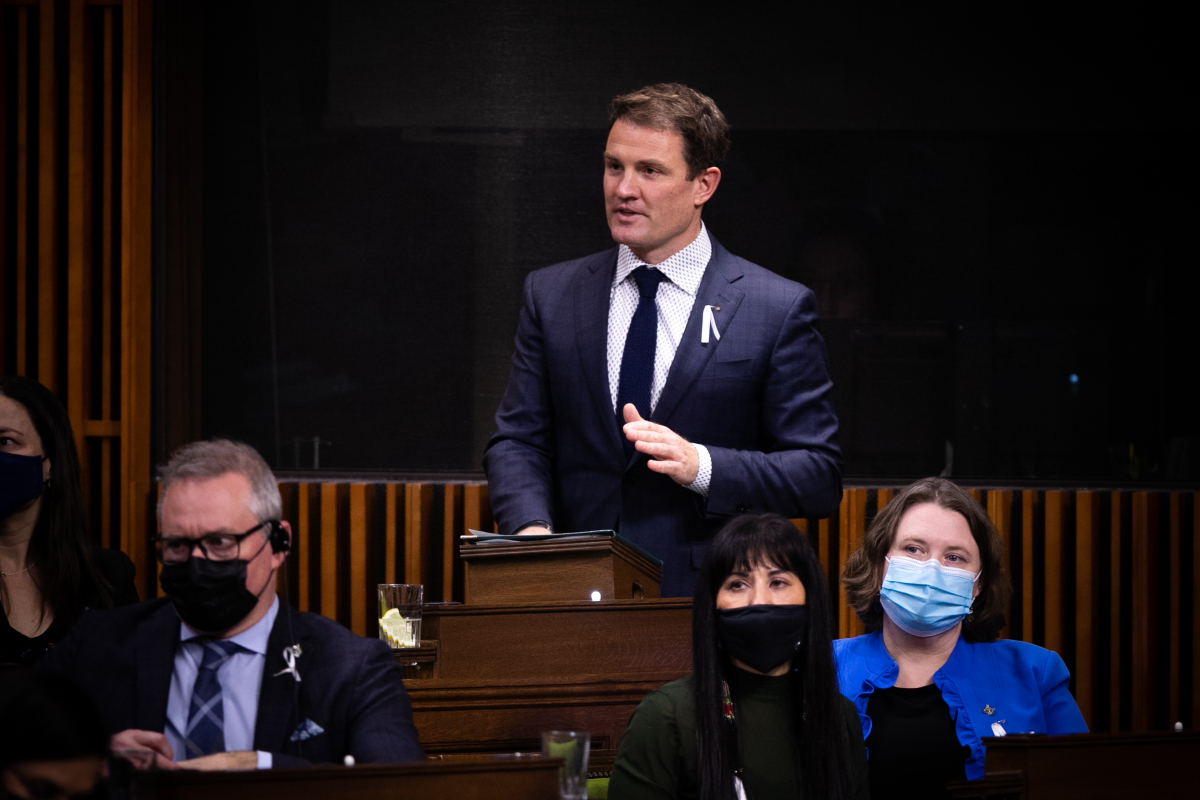 A man in a suit and tie stands in Canada's House of Commons, addressing his Parliamentary colleagues mid-sentence.