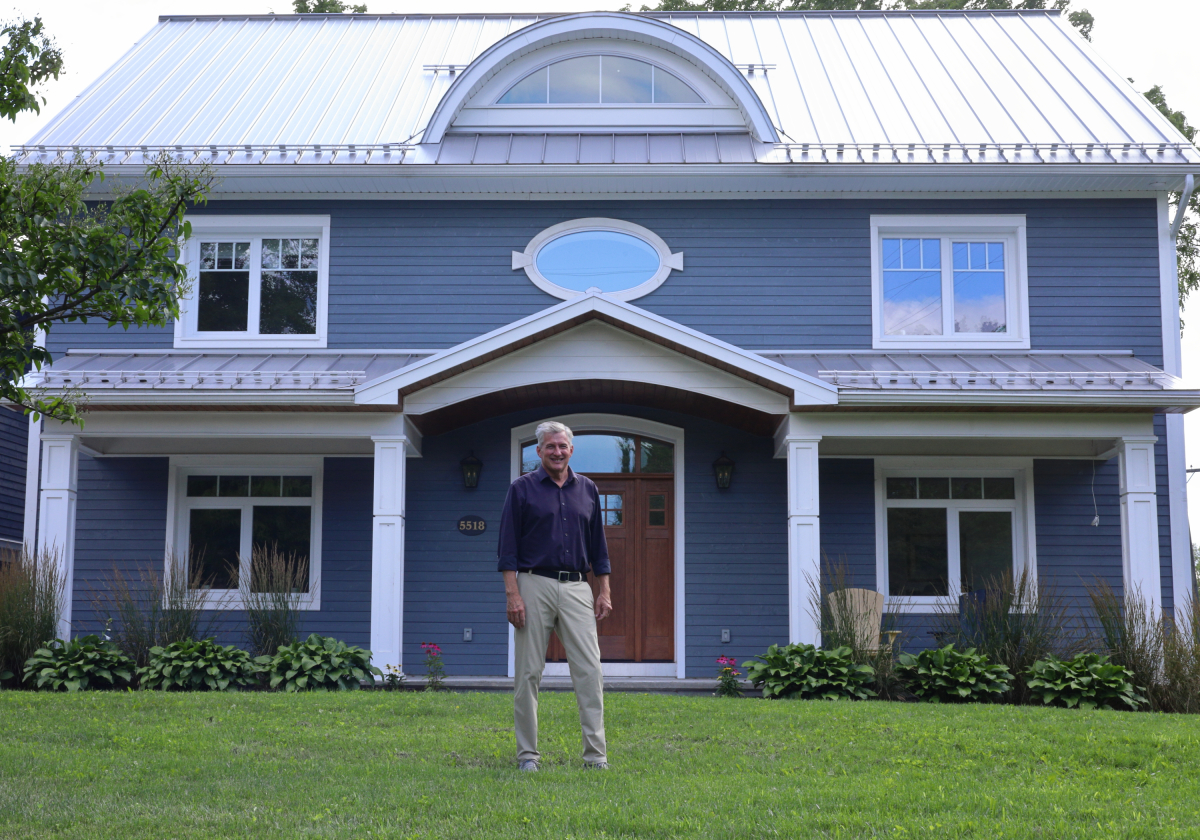 A man in a long sleeve collared shirt stands in front of a dark blue house on a bright, August afternoon