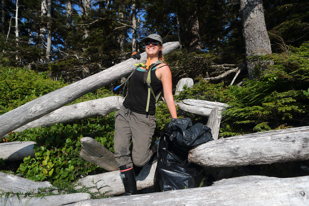 a woman holds up a piece of driftwood while collecting marine debris on a beach