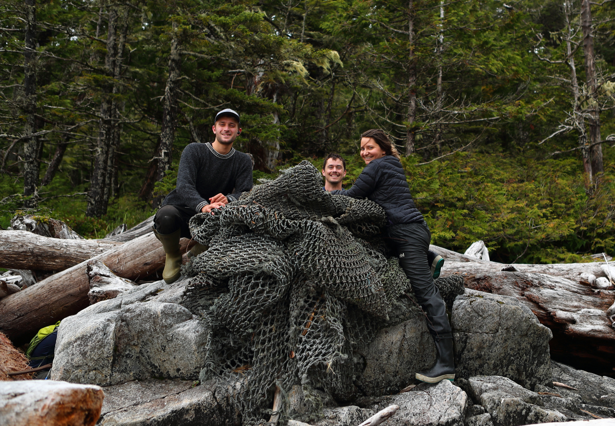 Three people pose with a large grey fishing net they removed from underneath rocks and driftwood on B.C.'s central coast.