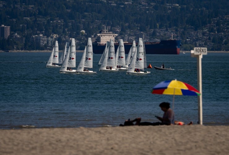 A person sits on the beach under a colourful umbrella as sail boats pass in Vancouver, B.C., on Sunday, July 7, 2024. Photo by The Canadian Press/Ethan Cairns